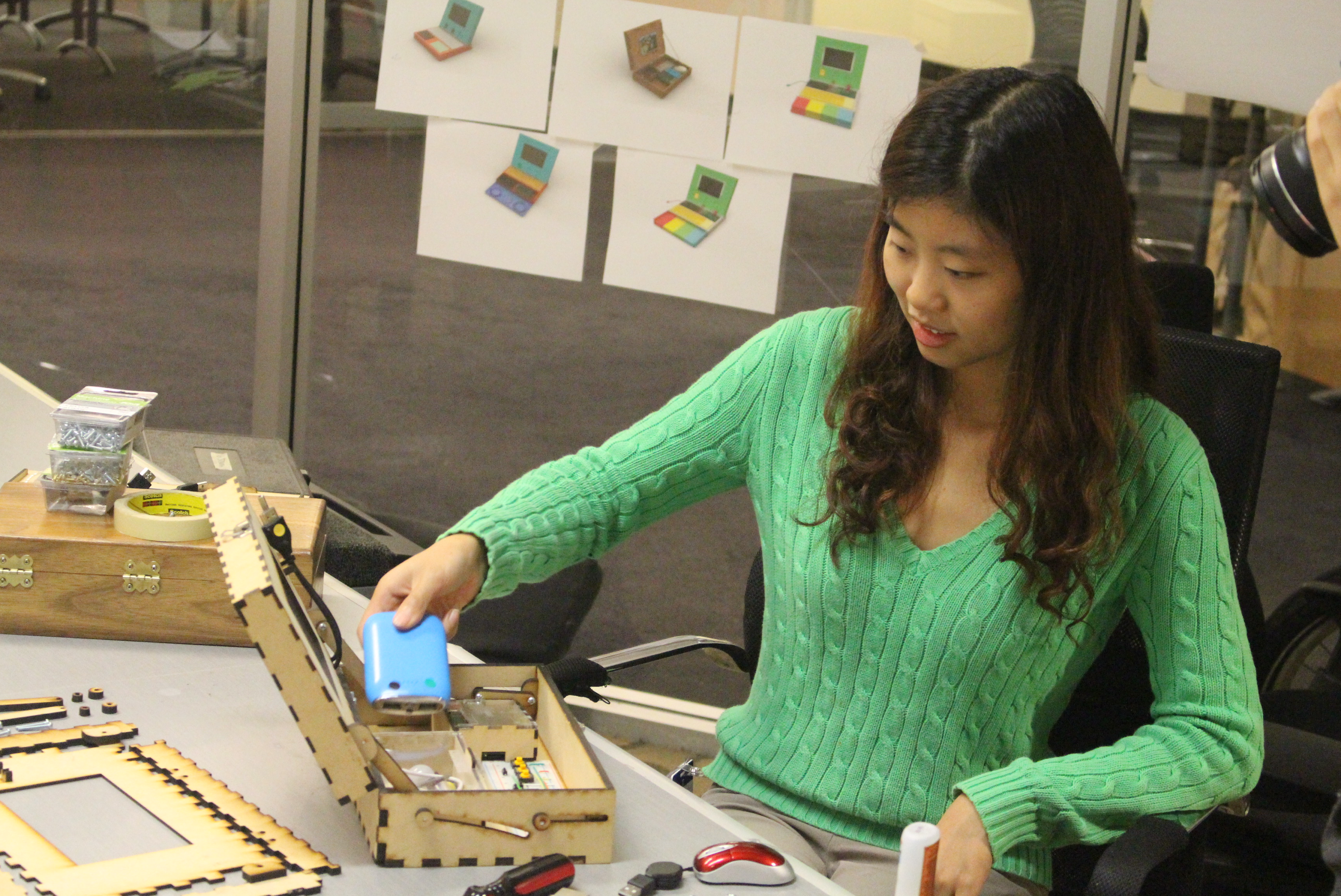Mary assembling a Piper prototype at her desk, color concept renderings pinned behind her